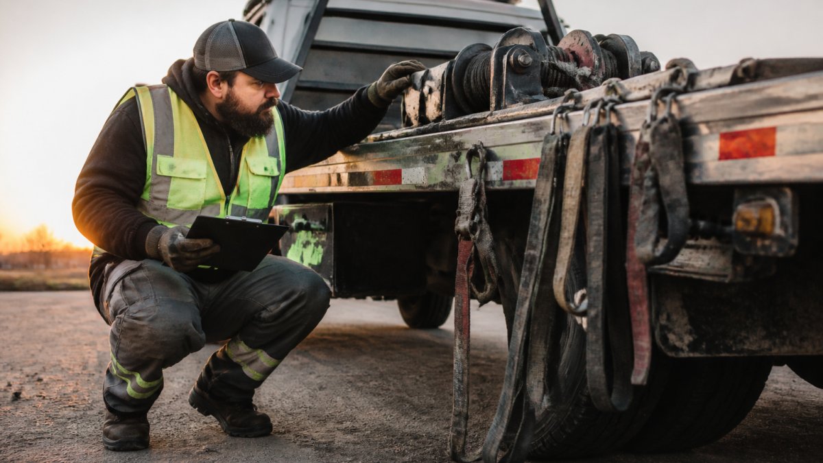A tow truck operator in a safety vest crouching beside a flatbed truck at dawn, inspecting the winch cable and straps with a clipboard in hand