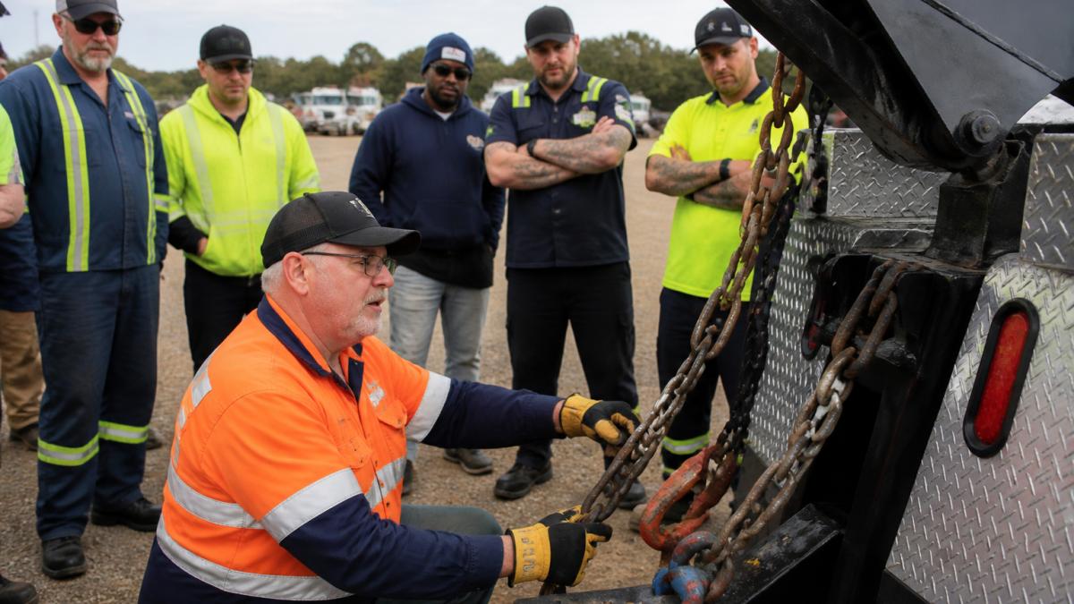 Professional instructor demonstrating proper rigging technique with heavy chains and vehicle equipment to a group of towing operators in an outdoor training yard with natural daylight